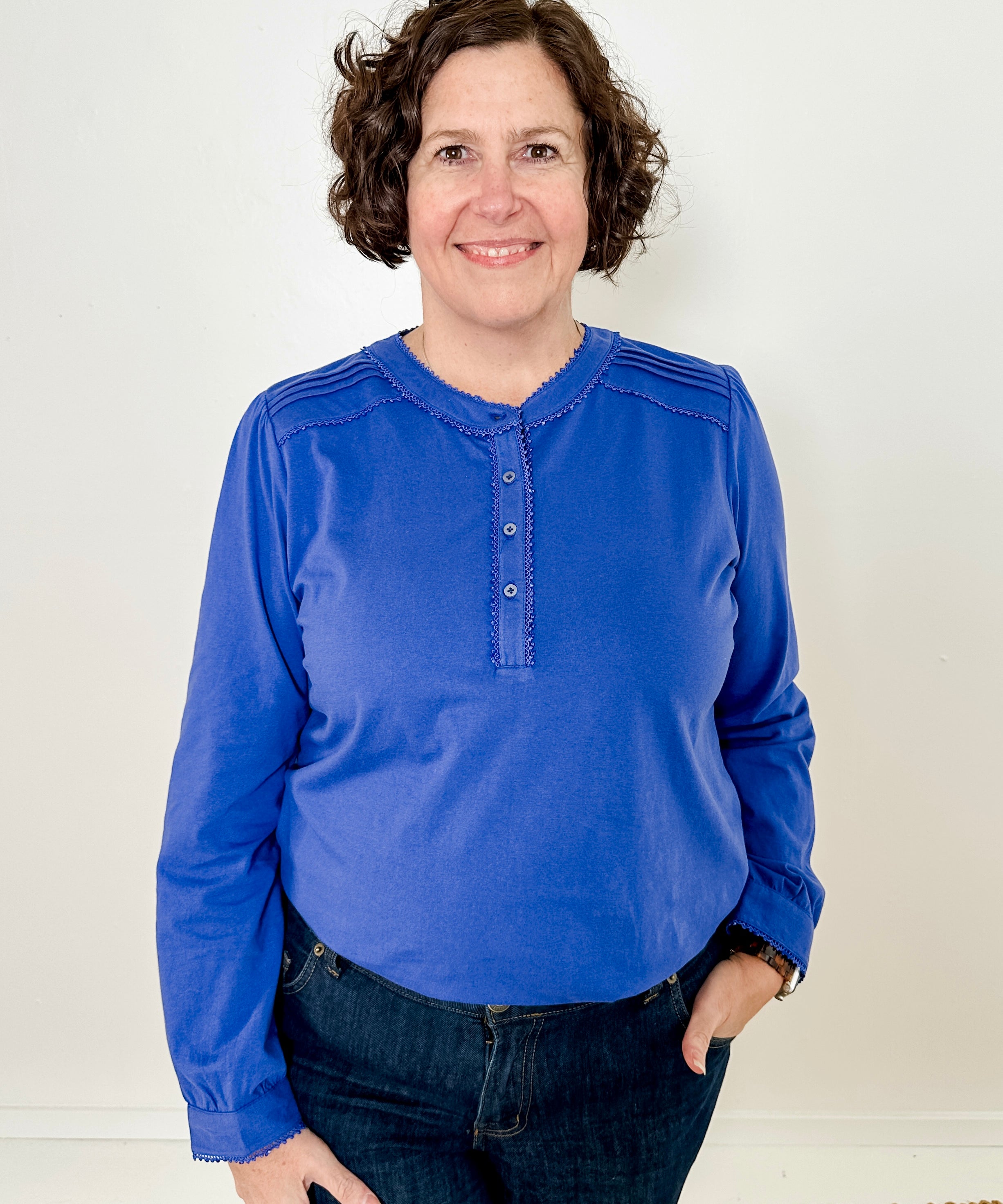 Women's knit blouse with lace detail, made from 100% cotton jersey surplus fabric. Available in sapphire. Features a collarless band neckline, matching button-front placket, delicate knit lace on shoulders, collar, cuffs, and placket, keyhole buttoned cuffs, and side slits. Medium-weight fabric with light stretch for a straight fit. Available in sizes XS-3X.