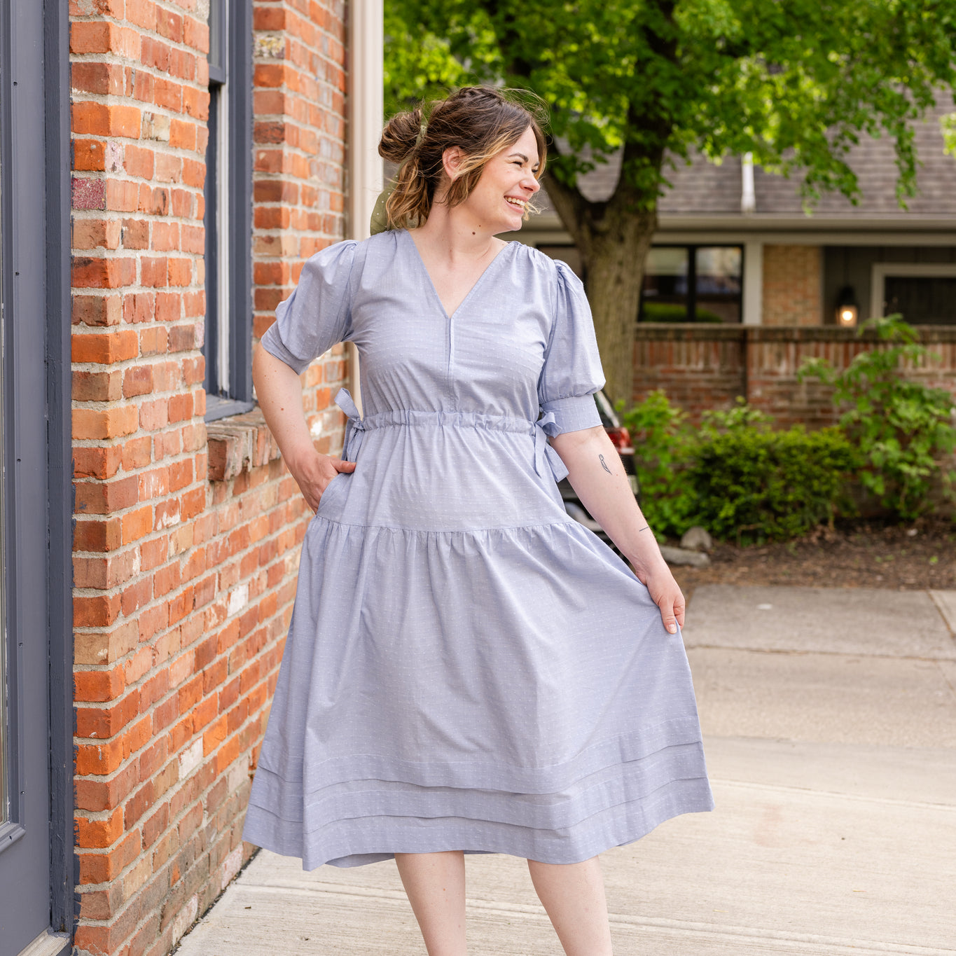 Elegant midi-length dress in surplus 100% cotton with puff sleeves and a unique V-neckline. Features ruching along the bodice, shoulders, and skirt, plus drawstring bows at the waist for a flattering fit. Designed with hidden side pockets, elbow-length keyhole cuff sleeves, and a box-pleated hem. Finished with French seams and a hidden decorative placket.