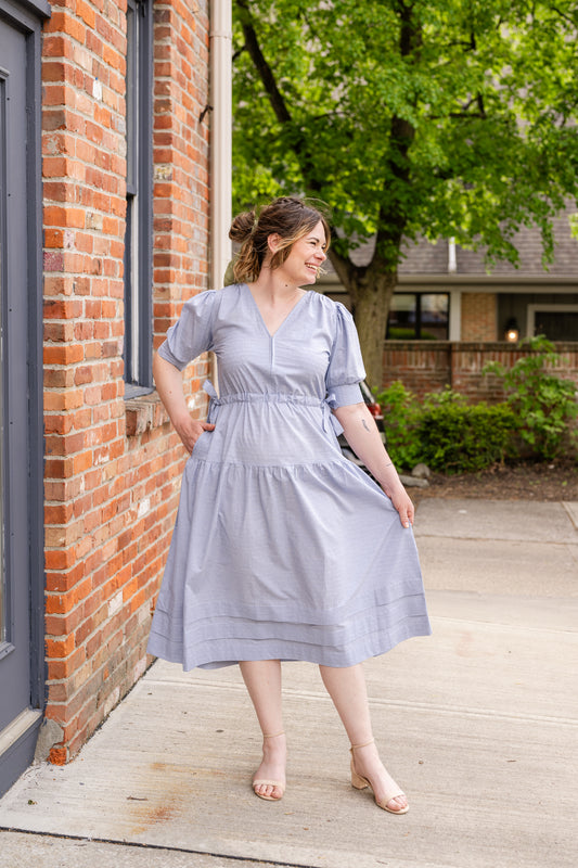 Elegant midi-length dress in surplus 100% cotton with puff sleeves and a unique V-neckline. Features ruching along the bodice, shoulders, and skirt, plus drawstring bows at the waist for a flattering fit. Designed with hidden side pockets, elbow-length keyhole cuff sleeves, and a box-pleated hem. Finished with French seams and a hidden decorative placket.