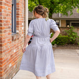 Elegant midi-length dress in surplus 100% cotton with puff sleeves and a unique V-neckline. Features ruching along the bodice, shoulders, and skirt, plus drawstring bows at the waist for a flattering fit. Designed with hidden side pockets, elbow-length keyhole cuff sleeves, and a box-pleated hem. Finished with French seams and a hidden decorative placket.