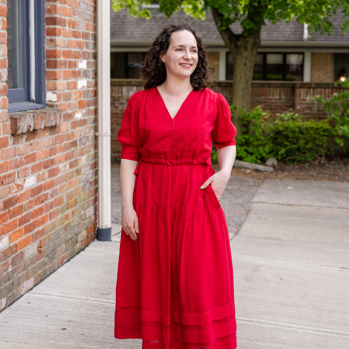 Elegant midi-length dress in surplus 100% cotton with puff sleeves and a unique V-neckline. Features ruching along the bodice, shoulders, and skirt, plus drawstring bows at the waist for a flattering fit. Designed with hidden side pockets, elbow-length keyhole cuff sleeves, and a box-pleated hem. Finished with French seams and a hidden decorative placket.