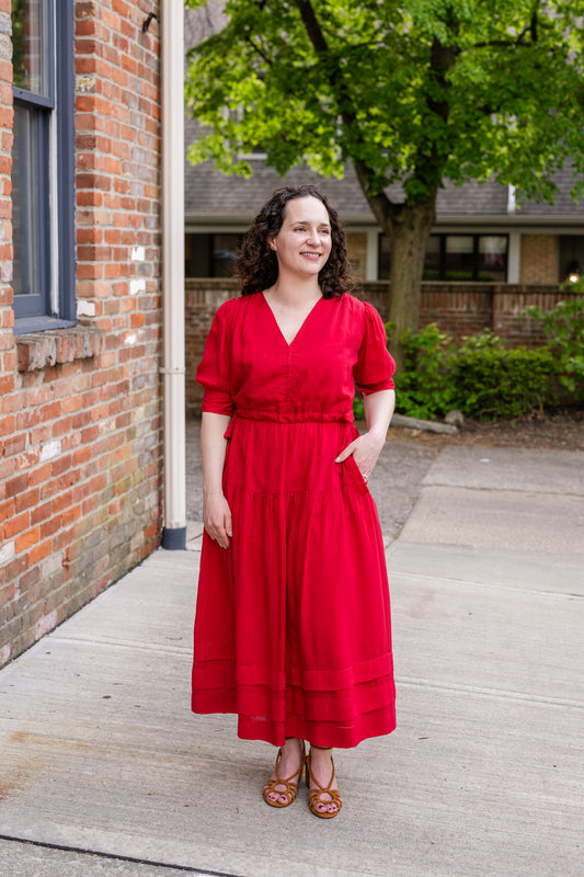 Elegant midi-length dress in surplus 100% cotton with puff sleeves and a unique V-neckline. Features ruching along the bodice, shoulders, and skirt, plus drawstring bows at the waist for a flattering fit. Designed with hidden side pockets, elbow-length keyhole cuff sleeves, and a box-pleated hem. Finished with French seams and a hidden decorative placket.
