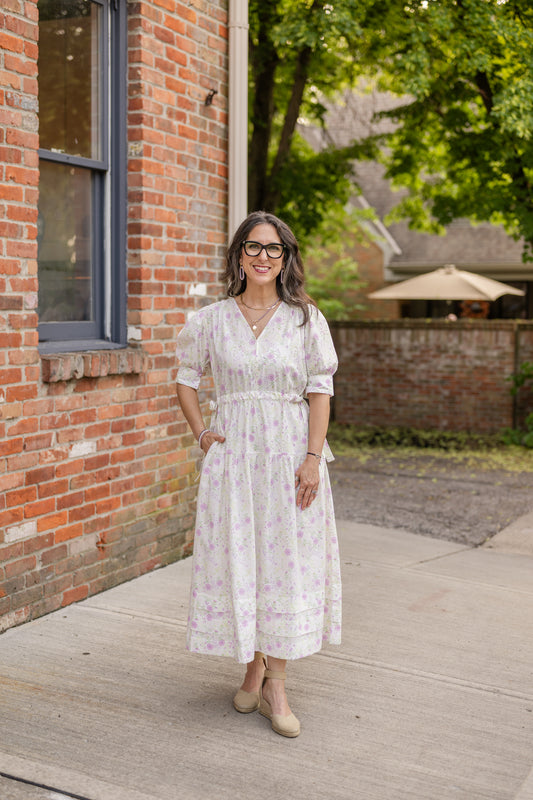 Elegant midi-length dress in surplus 100% cotton with puff sleeves and a unique V-neckline. Features ruching along the bodice, shoulders, and skirt, plus drawstring bows at the waist for a flattering fit. Designed with hidden side pockets, elbow-length keyhole cuff sleeves, and a box-pleated hem. Finished with French seams and a hidden decorative placket.